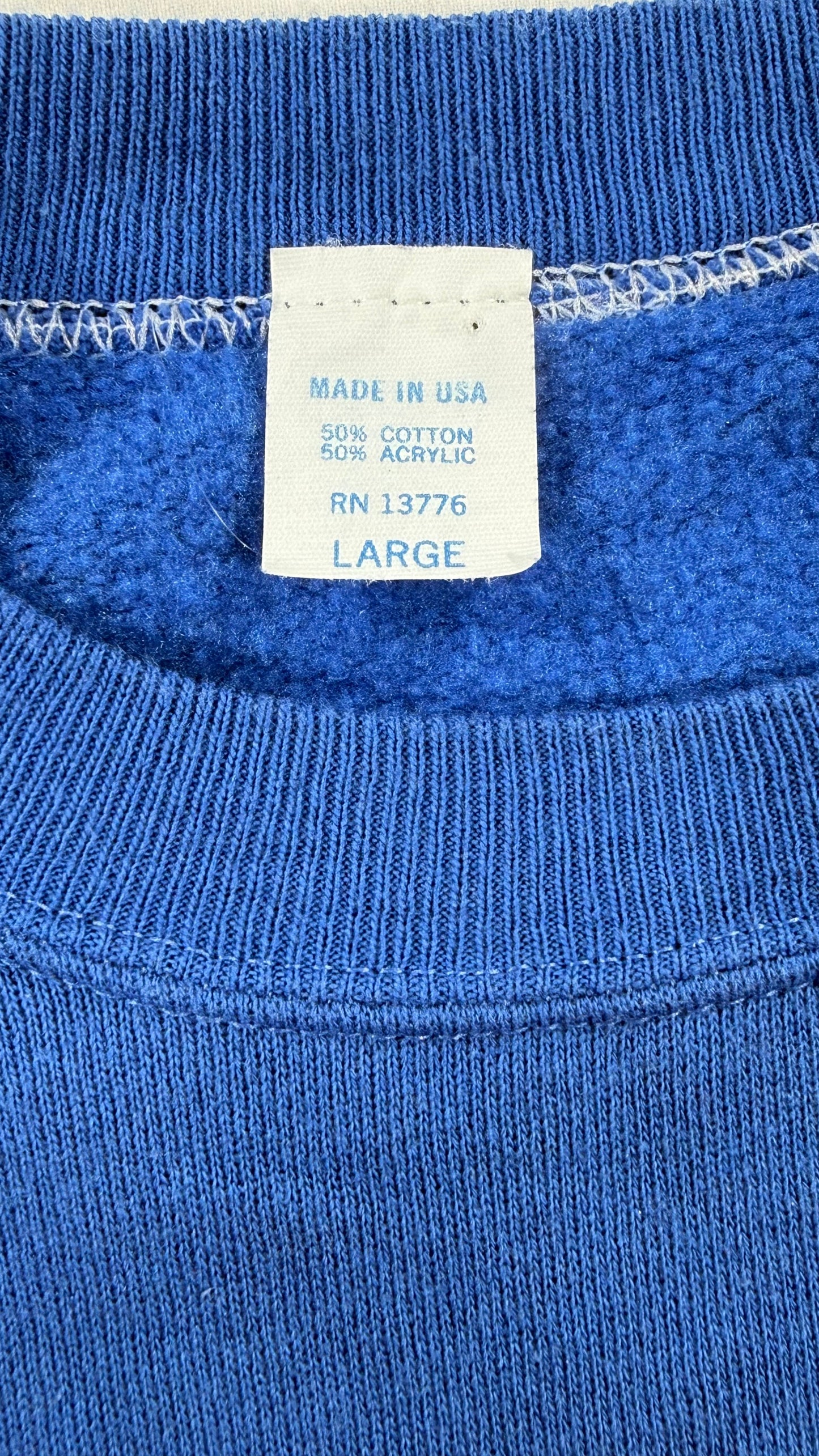 Close-up of the inside collar of a blue crewneck sweatshirt, showing a white sewn-in size label that reads 'MADE IN USA 50% COTTON 50% ACRYLIC RN 13776 LARGE'.
